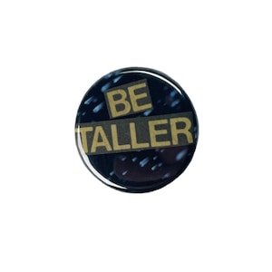 Puede incluir: Un botón redondo negro con las palabras "BE TALLER" en color beige claro. El botón tiene un acabado brillante y un fondo azul oscuro moteado.