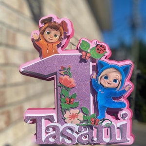 Könnte beinhalten: Ein mehrschichtiger, glitzernder rosa- und lilafarbener Tortenaufsatz mit der Zahl eins und dem Namen "Tasani". Der Aufsatz zeigt Zeichentrickfiguren in orangefarbenen und blauen Outfits sowie Blumen- und Schmetterlingsakzente. Ein Marienkäfer ist ebenfalls vorhanden.
