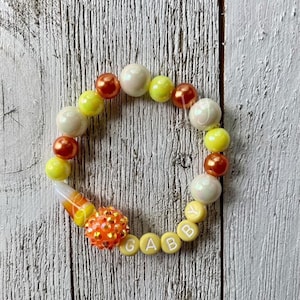 May include: A beaded bracelet with a candy corn theme. The bracelet features a mix of white, yellow, and orange beads, including a candy corn charm and beads that spell out the name "GABBY".