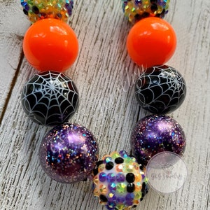 May include: A Halloween-themed necklace featuring a variety of beads. The necklace includes orange, black spiderweb, and purple glitter beads, along with sparkling, multi-colored rhinestone beads. The necklace is strung on a purple cord.