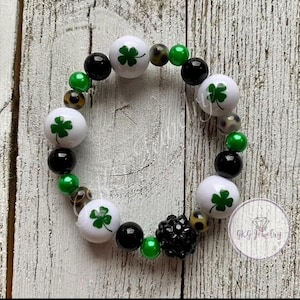 Könnte beinhalten: Ein Perlenarmband mit weißen Perlen mit grünen Kleeblättern, schwarzen Perlen, grünen Perlen und gemusterten Perlen. Eine schwarze, strukturierte Perle ist ebenfalls enthalten. Das Armband liegt auf einer hellen Holzoberfläche.