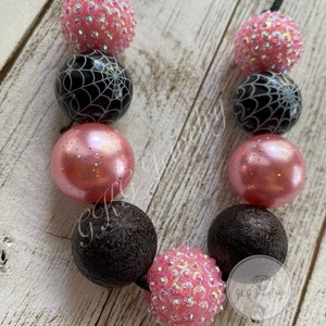May include: A beaded necklace featuring alternating pink and black beads. The pink beads have a glittery or textured appearance, while some black beads display a white spiderweb design. The necklace is strung on a black cord.