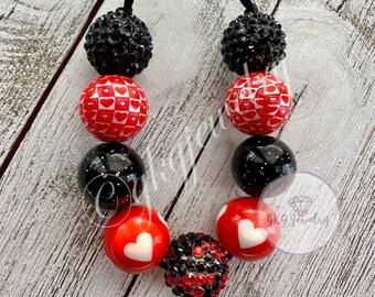 May include: A necklace featuring a variety of beads in red, black, and white. The beads include heart patterns, sparkling black beads, and beads with white hearts. The necklace is strung on a black cord.