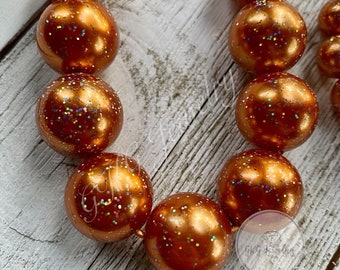 May include: A necklace featuring large, round beads in a metallic copper color with iridescent glitter. The beads are strung on an orange cord, creating a bold and eye-catching accessory. The beads are approximately 1 inch in diameter.