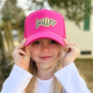 May include: A pink trucker hat with a gold "Willow" patch on the front. The hat is being worn by a young person with long blonde hair.