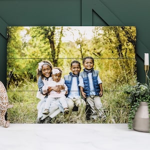 May include: A wood print featuring a photo of four children in a forest setting. The children are smiling and looking at the camera. The print is made of natural wood and has a rustic look.