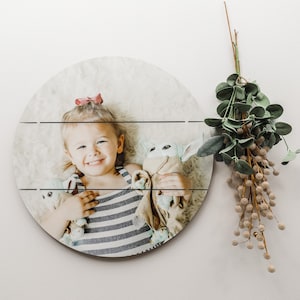 May include: A round wooden sign with a photo of a young girl smiling and holding a stuffed toy. The sign has a whitewashed wood background and is perfect for displaying a child's photo.