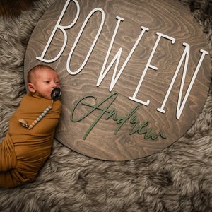 May include: A newborn baby boy swaddled in a brown blanket lies on a faux fur rug. A wooden sign with the name "BOWEN Andrew" is behind the baby.