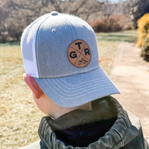 May include: A gray and white mesh trucker hat with a brown leather patch featuring the letters "TGR" and a mountain range design.