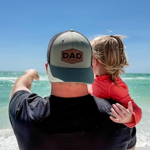 May include: A person wearing a green and black baseball cap with a brown leather patch that says "DAD est. 2023" is holding a young child in their arms. They are standing on a beach with the ocean in the background.