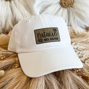May include: White baseball cap with a brown leather patch that says "Natalie BIG SISTER" in black lettering.