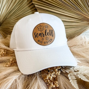 May include: White baseball cap with a brown leather patch that says "Scarlett Big Sister" with small flower designs.