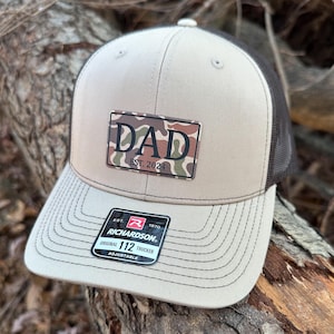 May include: A beige and black trucker hat with a camouflage patch that says "DAD EST. 2024". The hat has a black mesh back and a brown leather adjustable strap with a red and white logo that says "Richardson 112 Original Trucker Adjustable".