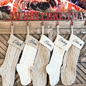 May include: Five cream-colored knitted stockings with black embroidered names: Dad, Mom, Eliana, Emmy, and Lexi with a paw print. The stockings are hanging from a red metal rack with the words "Merry Christmas" on it.