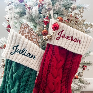 May include: Two Christmas stockings with names embroidered on them. The stocking on the left is green and has the name "Julian" embroidered on it. The stocking on the right is red and has the name "Jaxson" embroidered on it.