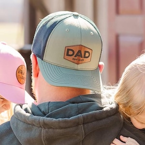 May include: A man wearing a green and blue baseball cap with a brown leather patch that says "DAD est. 2020" is holding two young girls. The girls are wearing pink and beige mesh baseball caps.