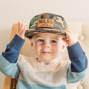 May include: A young child wearing a blue and white striped sweater, blue jeans, and a camouflage baseball cap with the word "MILES" on it. The child is sitting on a white tufted couch.