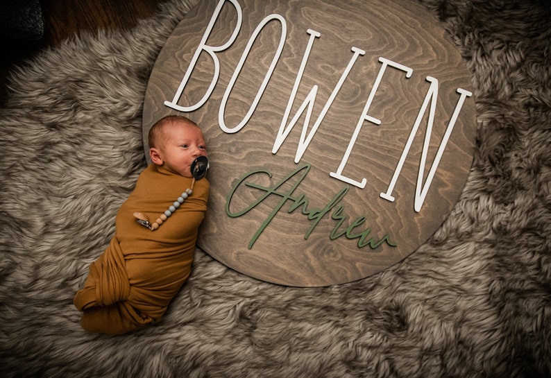 May include: A newborn baby boy swaddled in a brown blanket lies on a faux fur rug. A wooden sign with the name "BOWEN Andrew" is behind the baby.
