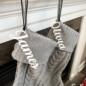 May include: Two gray knitted stockings with white personalized name tags. The name tags are attached to the stockings with black cords. The names on the tags are "Olivia" and "James".