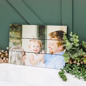 May include: A wood plank photo print featuring a family of three. A woman with blonde hair and glasses is holding a young girl with blonde hair. A man with brown hair is smiling at the girl. The photo is printed on a wood plank background.