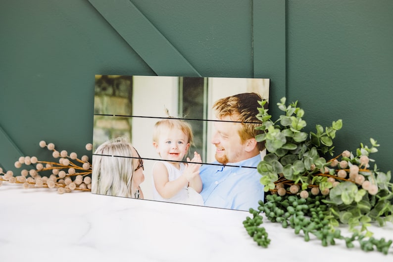 May include: A wood plank photo print featuring a family of three. A woman with blonde hair and glasses is holding a young girl with blonde hair. A man with brown hair is smiling at the girl. The photo is printed on a wood plank background.