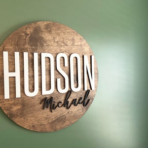 Op de afbeelding: Een rond houten bord met de naam "HUDSON Michael" in witte en zwarte letters.