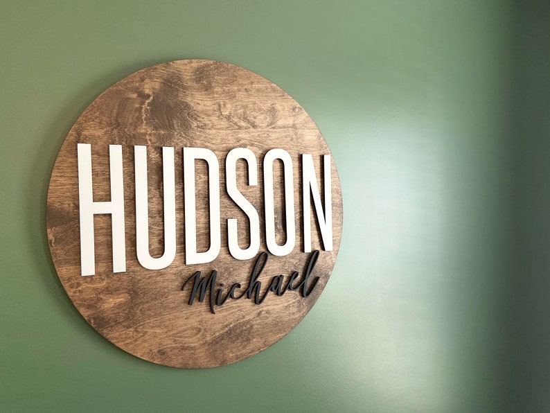Op de afbeelding: Een rond houten bord met de naam "HUDSON Michael" in witte en zwarte letters.