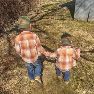 May include: Two children wearing plaid shirts and green baseball caps with their names, Benny and Logan, embroidered on the front. The children are walking in a grassy area.