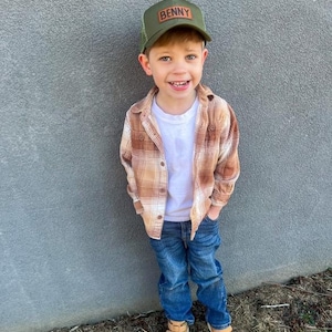 May include: A young boy wearing a green and brown plaid shirt, blue jeans, and a green mesh trucker hat with a brown leather patch that says "BENNY". He is smiling and looking at the camera.