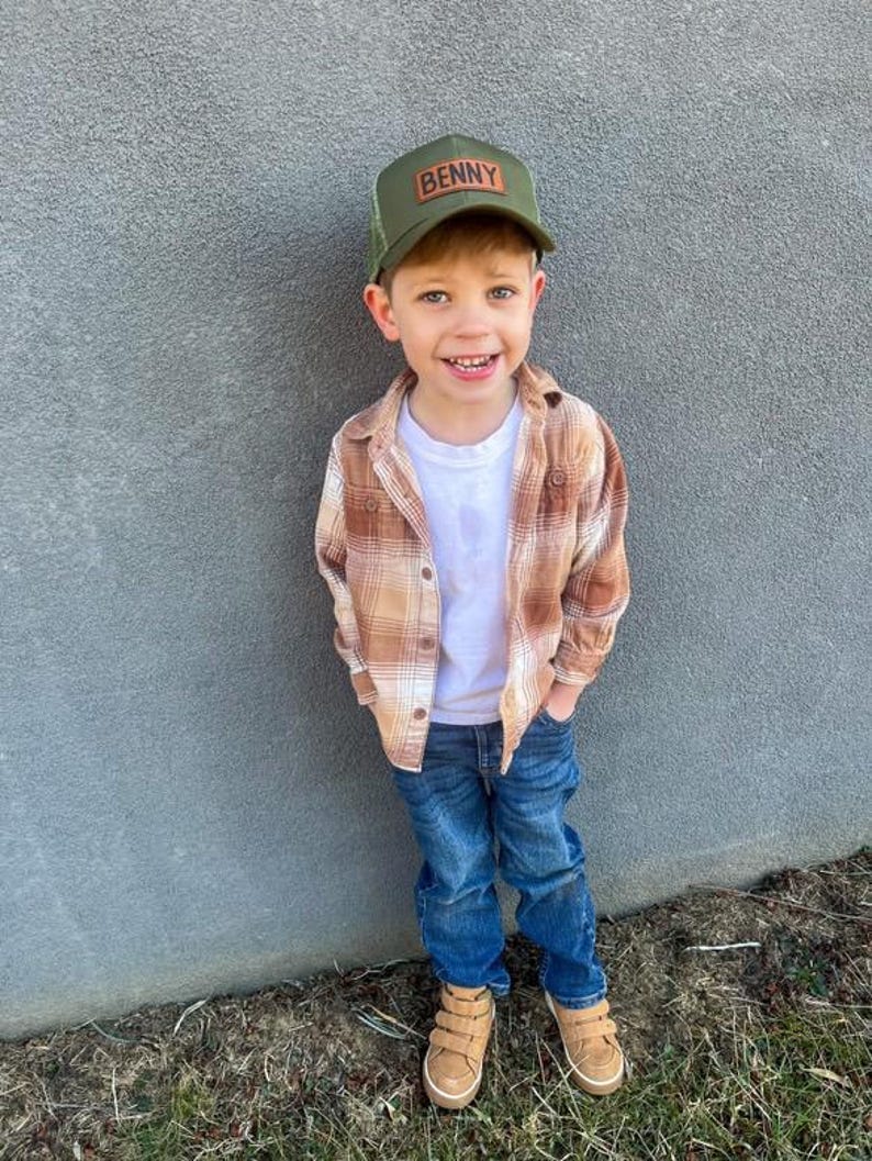 May include: A young boy wearing a green and brown plaid shirt, blue jeans, and a green mesh trucker hat with a brown leather patch that says "BENNY". He is smiling and looking at the camera.