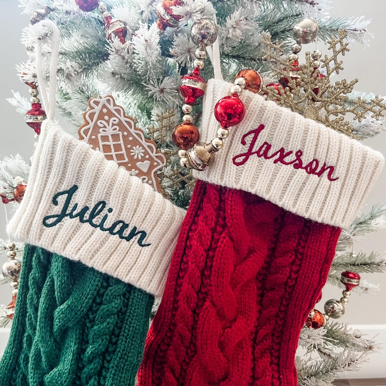 May include: Two Christmas stockings with names embroidered on them. The stocking on the left is green and has the name "Julian" embroidered on it. The stocking on the right is red and has the name "Jaxson" embroidered on it.