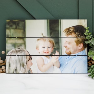May include: A wood plank photo display with a photo of a family of three. The photo shows a man holding a young child, while a woman looks on. The photo is printed on a wood plank background.