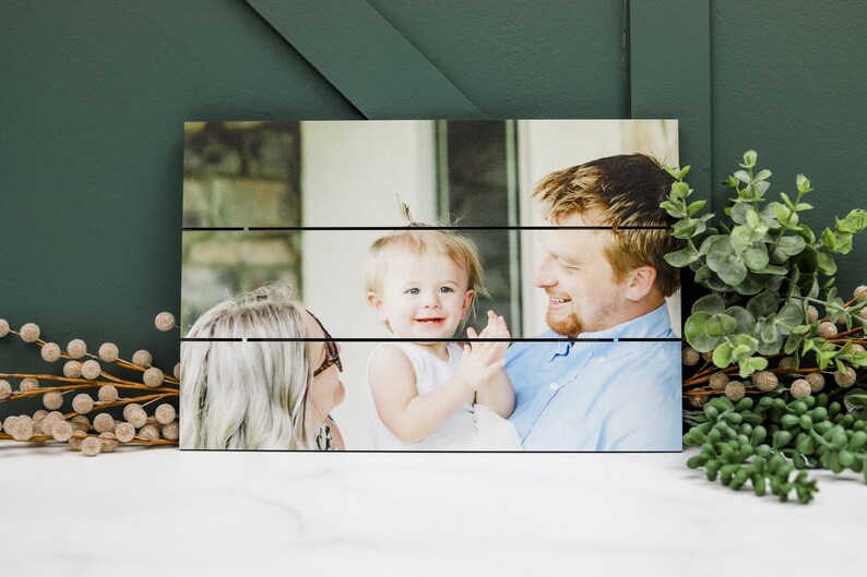 May include: A wood plank photo display with a photo of a family of three. The photo shows a man holding a young child, while a woman looks on. The photo is printed on a wood plank background.