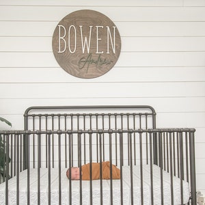 May include: A brown wooden round sign with the name "BOWEN" in white lettering and a cursive signature below. A baby is sleeping in a black metal crib with white bedding. There are potted plants on a shelf in the background.