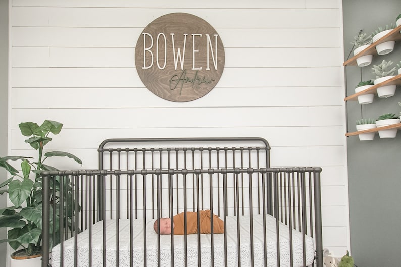 May include: A brown wooden round sign with the name "BOWEN" in white lettering and a cursive signature below. A baby is sleeping in a black metal crib with white bedding. There are potted plants on a shelf in the background.