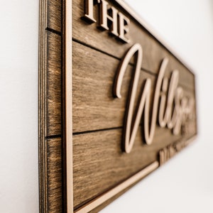 May include: A wooden sign with a brown stain and a light brown frame. The sign says "The Wilson Family" in a cursive font.