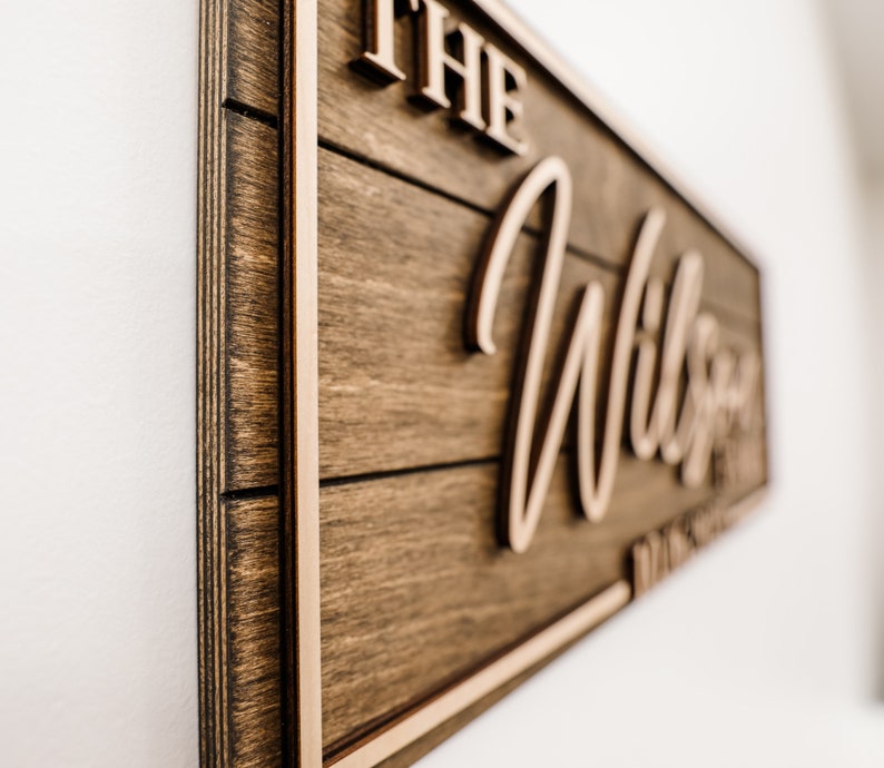 May include: A wooden sign with a brown stain and a light brown frame. The sign says "The Wilson Family" in a cursive font.