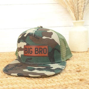 May include: Camouflage baseball cap with a brown leather patch that reads "BIG BRO". The cap features a green mesh back and a curved brim. The hat is designed with a classic trucker style.