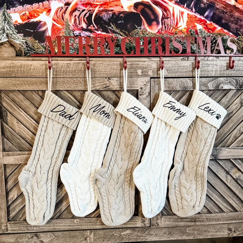 May include: Five cream-colored knitted stockings with black embroidered names: Dad, Mom, Eliana, Emmy, and Lexi with a paw print. The stockings are hanging from a red metal rack with the words "Merry Christmas" on it.