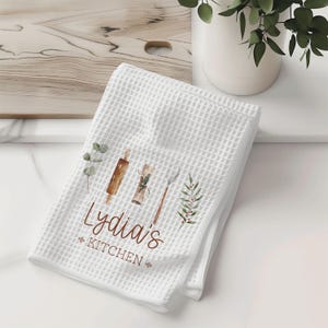 May include: White waffle weave kitchen towel with a brown and white watercolor design of kitchen utensils and greenery. The text "Lydia's Kitchen" is printed in brown.