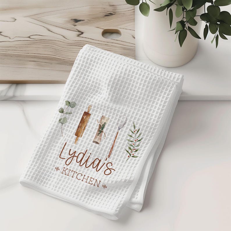 May include: White waffle weave kitchen towel with a brown and white watercolor design of kitchen utensils and greenery. The text "Lydia's Kitchen" is printed in brown.