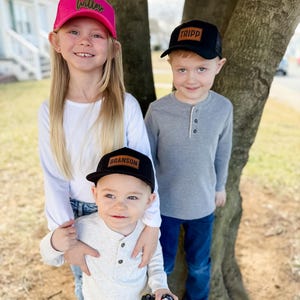 May include: Three children wearing black and pink trucker hats with personalized leather patches. The hats have mesh back panels. The children are standing in front of a tree.