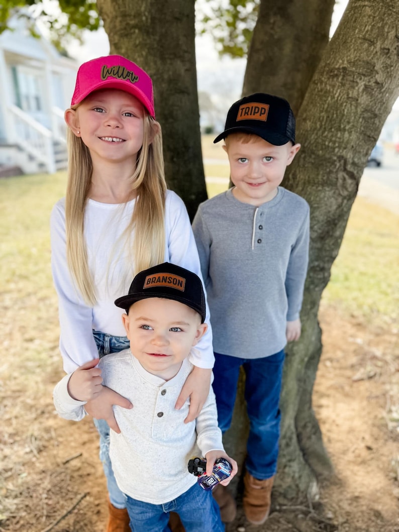 May include: Three children wearing black and pink trucker hats with personalized leather patches. The hats have mesh back panels. The children are standing in front of a tree.