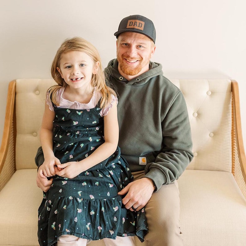 Puede incluir: Un hombre con una gorra negra con el texto "DAD" y una sudadera con capucha verde est&aacute; sentado con una ni&ntilde;a peque&ntilde;a con un vestido floral. Est&aacute;n sentados en una silla color crema con botones.
