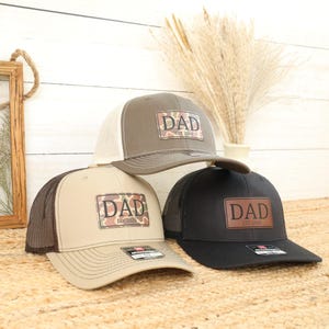 May include: Three trucker hats with the word "DAD" on the front. One hat is olive green with a white mesh back, another is tan with a brown mesh back, and the third is black with a black mesh back. Each hat has a different year.