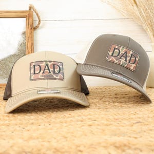 May include: Two trucker hats with a camouflage patch that reads "DAD" and the year of establishment. One hat is tan with a brown mesh back, and the other is brown with a white mesh back. The hats are on a woven surface.