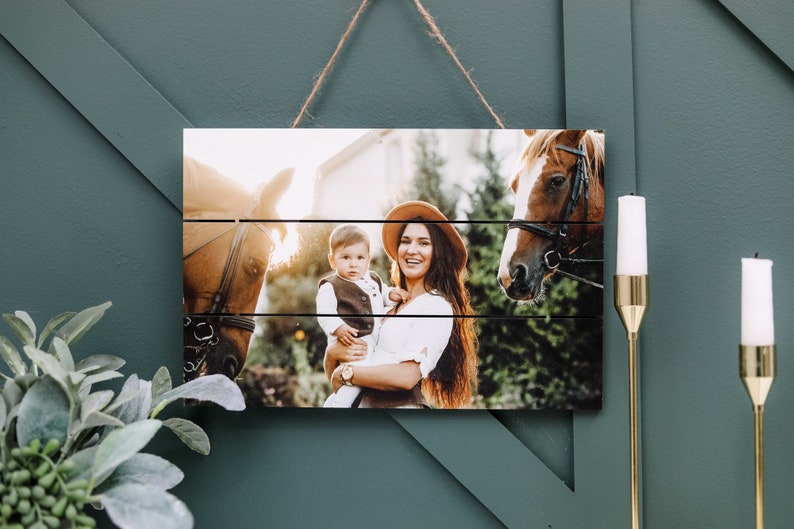 May include: A wooden photo print with a rustic look, featuring a woman holding a young child in front of two horses. The woman is wearing a brown hat and a white shirt. The child is wearing a vest and a white shirt. The horses are brown and white.