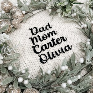 May include: A white knitted background with a green wreath with white berries.  The wreath surrounds four black words: Dad, Mom, Carter, Olivia.