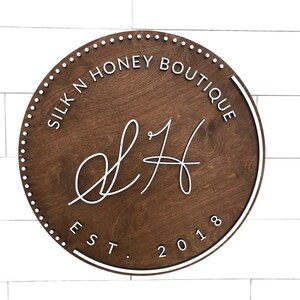 Op de afbeelding: Een rond houten bord met een witte rand en witte letters. Het bord zegt "Silk N Honey Boutique" en "Est. 2018". De letters "S&H" zijn in een cursief schrift.