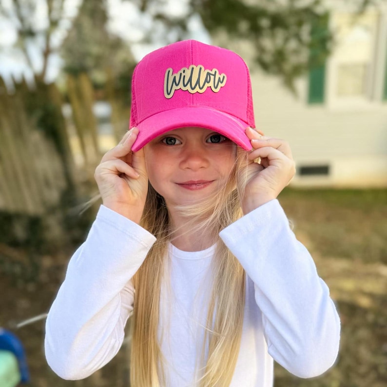 May include: A pink trucker hat with a gold "Willow" patch on the front. The hat is being worn by a young person with long blonde hair.